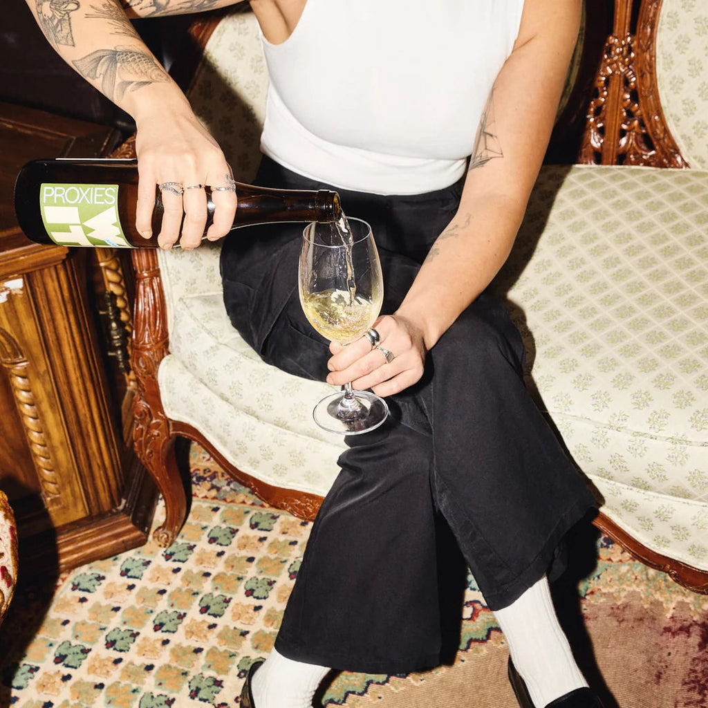 A person sitting on an ornate vintage sofa pours wine from a bottle labeled PROXIES into a glass, wearing a white tank top, black wide-leg pants, white socks, and black loafers in a decorated room.