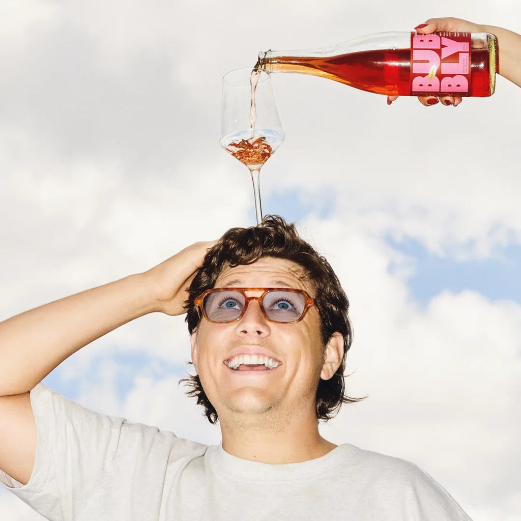A man wearing glasses looks up as a hand pours a pink drink from a bottle labeled 'Proxies Bubbly' into a glass balanced on his head, against a cloudy sky background.